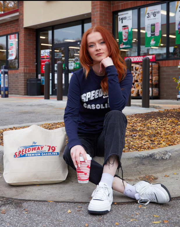 Woman sitting on a curb wearing a Speedway shirt with a Speedway tote bag next to her