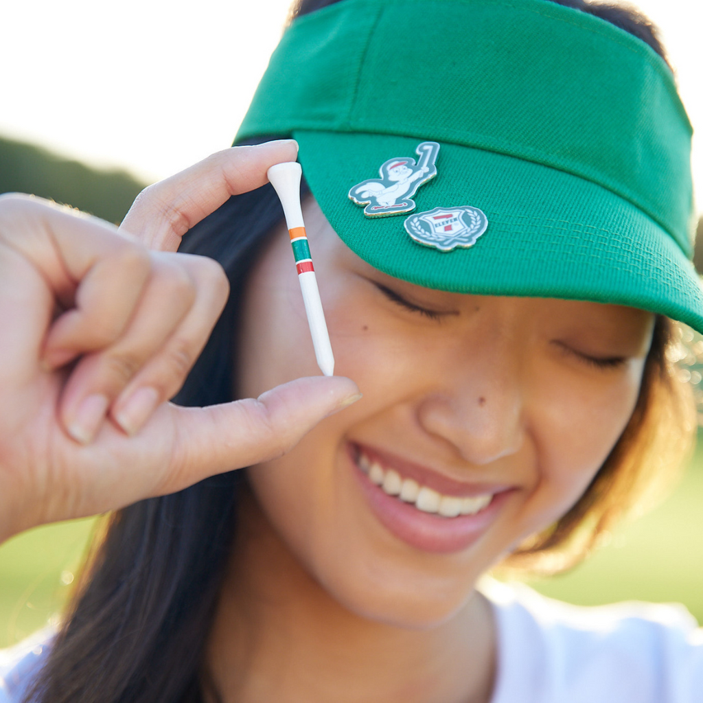 Woman holding a golf tee while wearing a visor with 7-Eleven enamel pins on it.